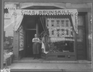 Man in a leather apron standing next to a saddle in the doorway of a shop with the sign Chas. Brunskill Harness and Leather Goods Horse Sundries.