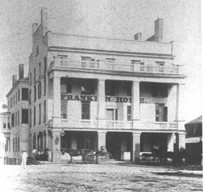 View of the front of a 24-story building with two balconies and four pillars labeled The Franklin Hotel. Several people and horse-drawn wagons stand outside.