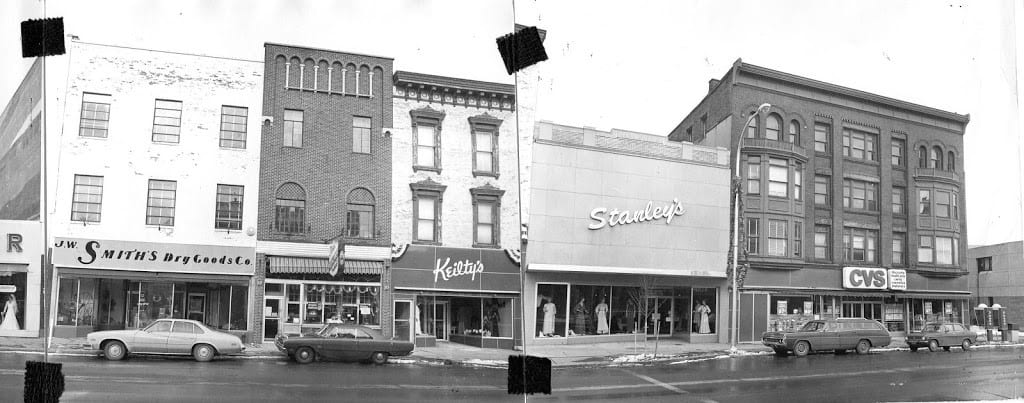 Two photos taped together of buildings. Shows 1970s cars parked out front of JW Smith's, Keilty's, Stanley's and CVS.