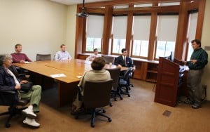 A group of people sitting around a conference table in a classroom listening to a man standing at a lecturn.