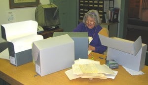 Older woman sitting at a table with archival boxes and piles of paper on it.