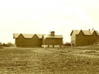Black and white view across bare fields towards five buildings including an Italianate house and several barns.