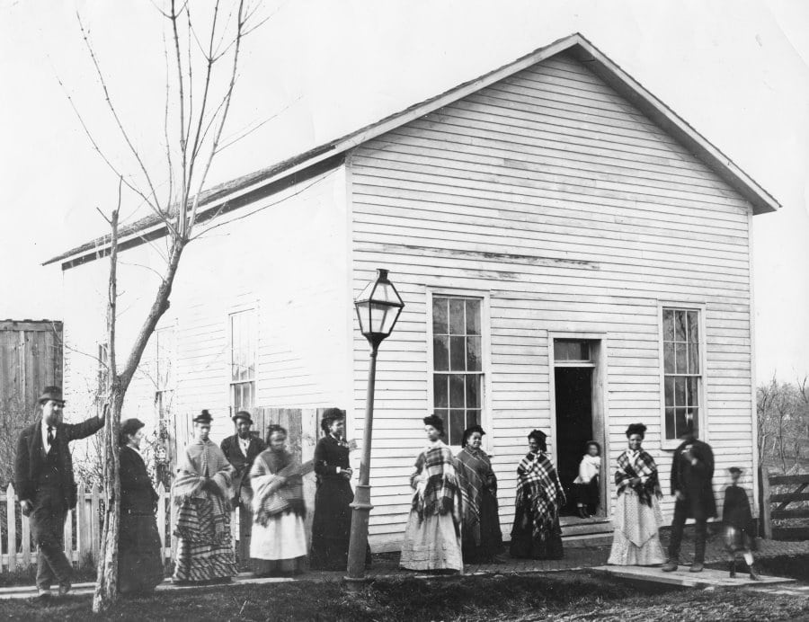 A group of mostly black men and women wearing shawls and hats stand on a sidewalk outside a white clapboard building with a lampost.