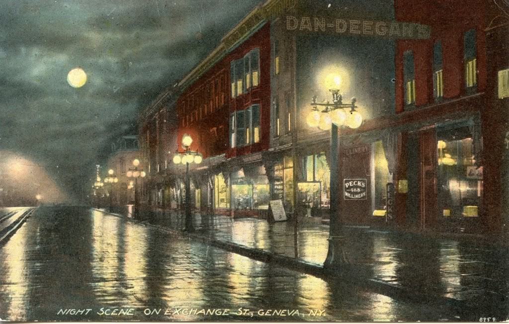 Postcard showing a commercial street of brick buildings with gas street lamps lit.