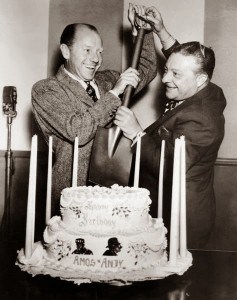 Two men in suits pretend to cut a 2-layer cake with a garden spade. The cake is surrounded by nine tapered candles and has "Happy Birthday Amos n Andy" on it in frosting along with the images of two black men.