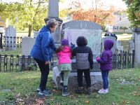 Three children and an adult standing in a cemetery in front of a gravestone doing a rubbing of the inscription.