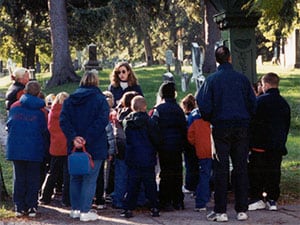 A woman and a group of children standing under a metal arch at the entrance of a cemetery.