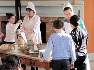 Women in old-fashioned dress churning butter and mixing batter with chldren.