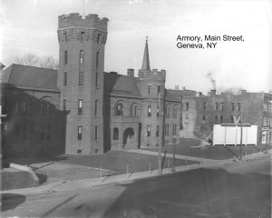 Black and white photo of a castle-like building on Main Street in Geneva. A cannon sits on the lawn next to a large white sign with a V at the top center over a Kiwanis logo.