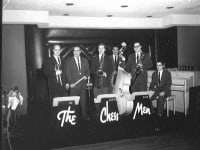 Six men standing with their instruments (trumpet, drumsticks, 2 saxaphones, an upright bass and a piano) behind stands that read The Chess Men.