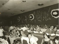 People sitting at tables and in booths at a club. The wall is decorated with the drawings of performers like the Four Aces and the Mills Brothers.