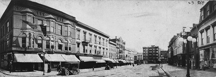 A panoramic view of a city block of brick buildings with awnings over storefronts and old-fashioned cars on the street.