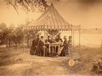 Seven people in old-fashioned dress sitting around a table set for tea under a tent looking out on a lake.