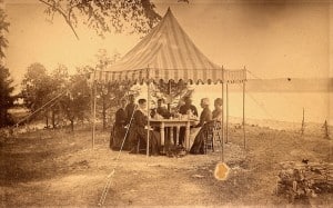 Seven people in old-fashioned dress sitting around a table set for tea under a tent looking out on a lake.