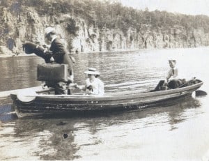 A young girl and man are siting in a small boat, while an older man with a suitcase and package gets out onto a dock.