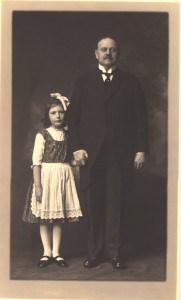 A little girl in a dress and apron standing holding hands with a man in a suit.