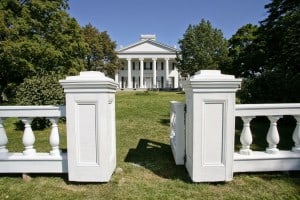 Looking at an elegant 6-columned Greek revival house across an expanse of lawn from just outside a low white gate.