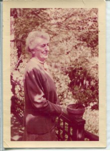 A faded color photograph of an older woman with white, short hair dressed in a suit and standing at a railing in a garden.