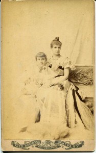 Portrait studio image of a middle-aged woman sitting on a bench with a young woman leaning against her. Both are in long formal gowns of the 1890s.