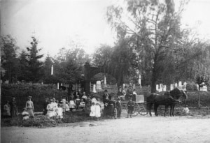 Large group of people and a horse-drawn buggy are gathered in front of a cemetery entrance.