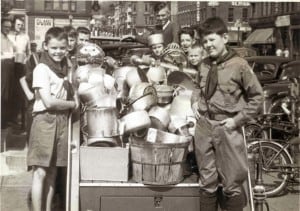 Boys in scout uniforms standing around a pile of scrap metal in a downtown street.