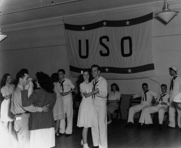 Sailors and women dancing in front of a USO banner.