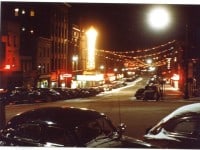 Veiw past old-fashioned cars down a lit street at night. A large sign for the Schine theater is visible.