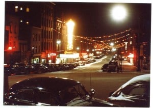 View past old-fashioned cars down a lit street at night. A large sign for the Schine theater is visible.