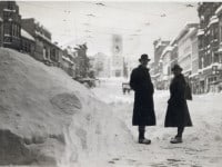 A man and woman standing next to high snowbanks in a street with commercial buildings on both sides and a church at the end.
