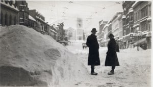 A man and woman standing next to high snowbanks in a street with commercial buildings on both sides and a church at the end.