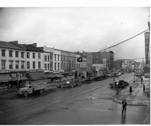 A view down Seneca Street showing 1940s cars parked on the side and wreaths hanging from garlands stretched between buildings across the road.