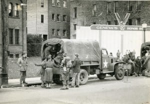 Soldiers and other people outside a building standing near a canvas covered truck full of men.