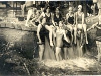 Girls in 1920s bathing suits and caps sitting on the edge of a small waterfall.