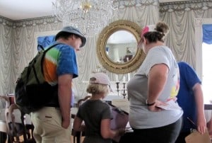 A man wearing a hat and backpack, a child and a woman stand around a table looking down at something on it in a fancy dining room with a crystal chandelier.