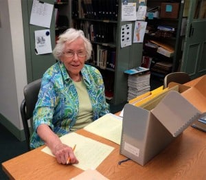 Colored photograph of an elderly woman working at a table in a research room
