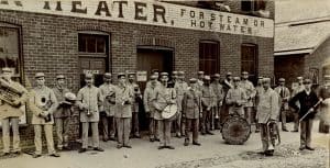 A group of men in band uniforms carrying horns and drums and standing in front of a brick building.
