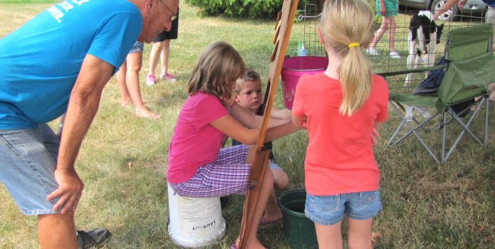 three children milk a fake cow with a man looking on