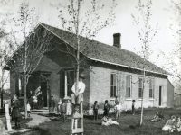 A one-room brick schoolhouse with two teachers and a group of children sitting and standing in the yard.