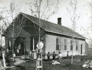 A one-room brick schoolhouse with two teachers and a group of children sitting and standing in the yard.