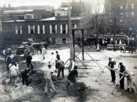 A large open city lot with a group of about 20 men shoveling and moving dirt or placing playground equipment while a crowd of people watches.