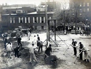 A large open city lot with a group of about 20 men shoveling and moving dirt or placing playground equipment while a crowd of people watches them construct the first Geneva playground.