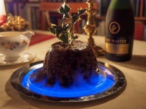A brown, rounded pudding on a silver tray with a holly sprig inserted in the top and blue flames around the circumference.