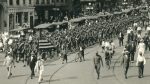 Parade of men in army uniforms accompanied by civilians walking and biking on road with trolley tracks.