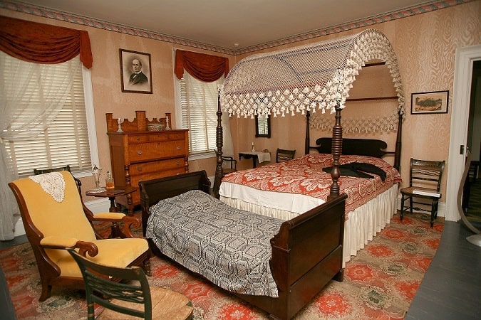 A room with a four-poster bed with a lace canopy, a red coverlet and a twin bed at its foot.