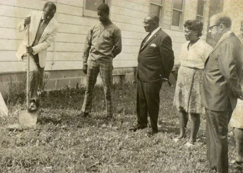 Three men and a woman watch a man dig with a shovel just outside a building.