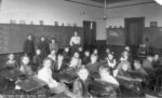 26 students standing by a blackboard or sitting in old-fashioned desks in rows with a teacher looking on.