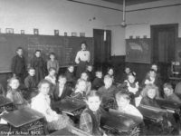 26 students standing by a blackboard or sitting in old-fashioned desks in rows with a teacher looking on.