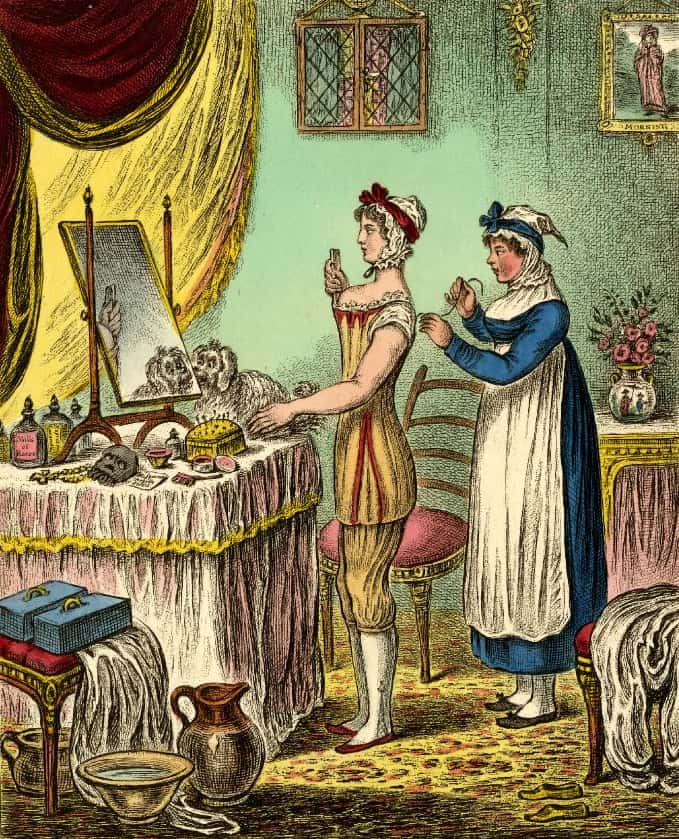 Illustration of a woman standing at a dressing table in undergarments getting laced into her corset by a maid.