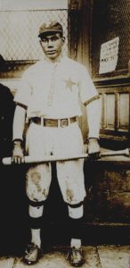 An African-American man in a baseball uniform and holding a bat in front of his thighs.
