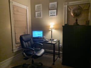 In between a window and a cupboard is an office chair and a table with a lamp and computer on it.